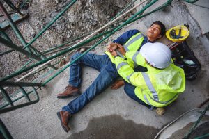 A male construction worker wearing a hi-vis jacket lying on a concrete floor underneath scaffolding on his back. His helmet has come off his head and is beside him. Another male construction worker is crouched beside him.