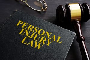 A black law book with the words 'personal injury law' on the front in gold. Beside the book is a pair of glasses and a black gavel, all on a black table. 