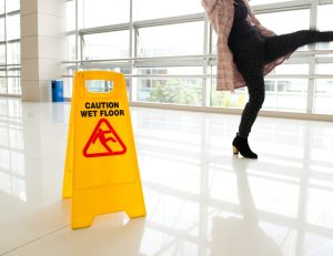 A yellow wet floor sign in a white hallway, in the background there is a woman with her leg up mid-slip.