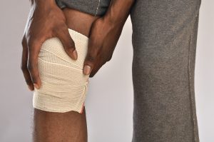 A close-up shot of a knee wrapped in a bandage. The injured man is holding his bandaged knee in pain.